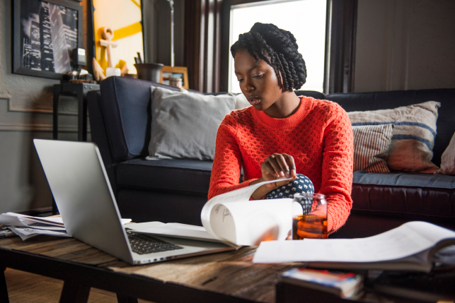 Woman seated on floor flipping through pages with laptop on coffee table