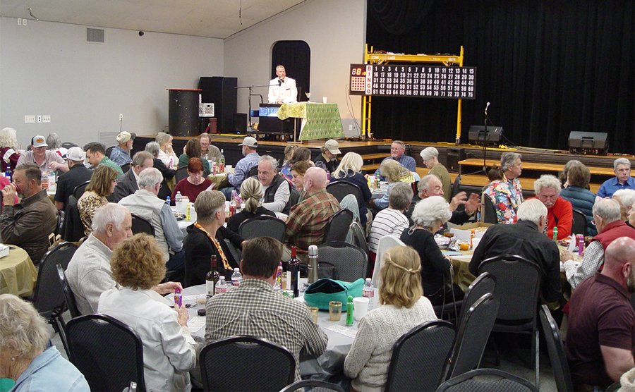 Man on a stage next to a large bingo board looking on while a large crowd of older folks sit around tables with bingo supplies.