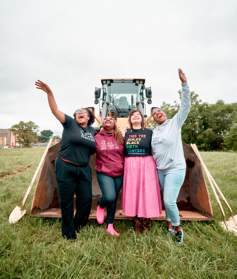 Four women stand arm-in-arm in front of a bulldozer to celebrate the groundbreaking of their new birthing center, Birth Detroit.