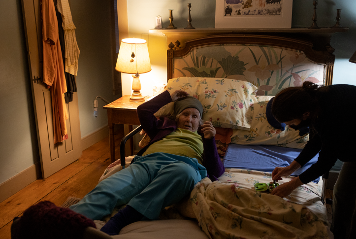 Older woman lies in bed while being helped by younger person